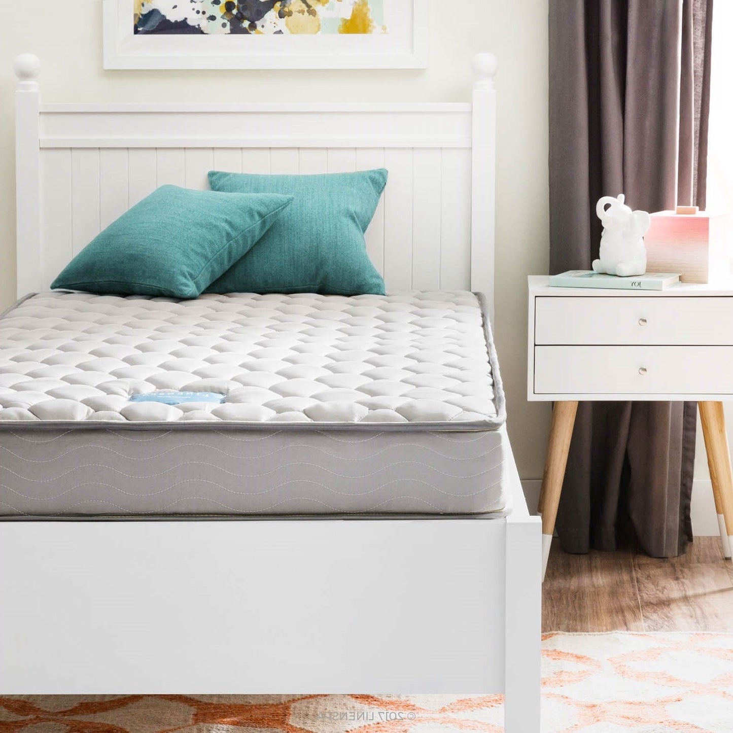 Gray mattress on a white bed frame with teal pillows in a bedroom setting.