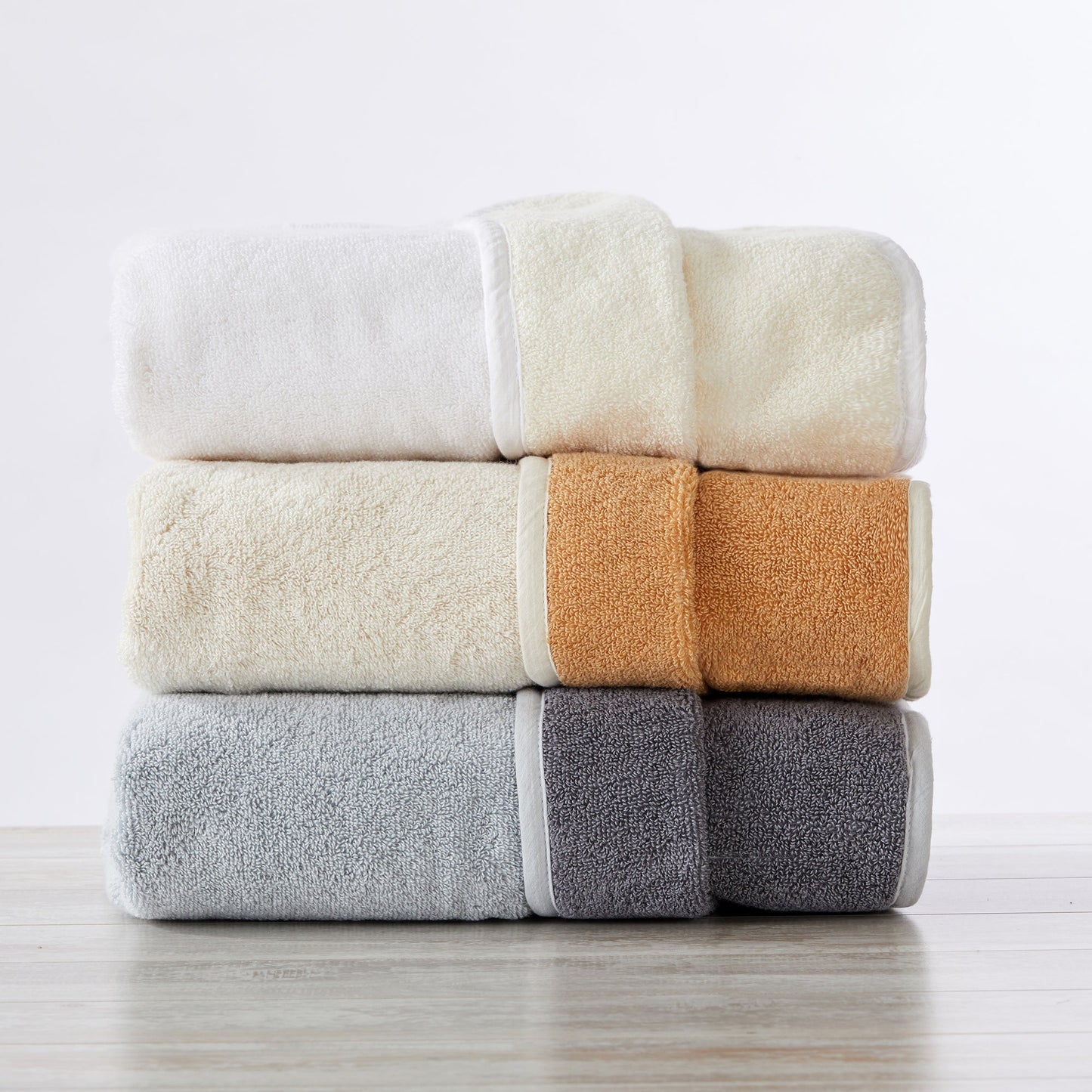 Stack of four folded towels in white, beige, orange, and gray on a light wooden surface.