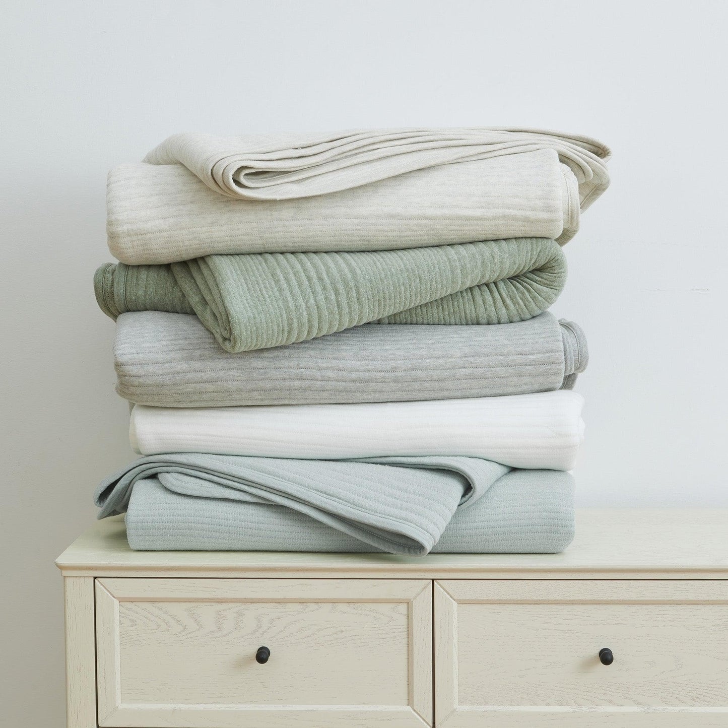 Stack of folded blankets in various shades of green and gray on a white dresser.