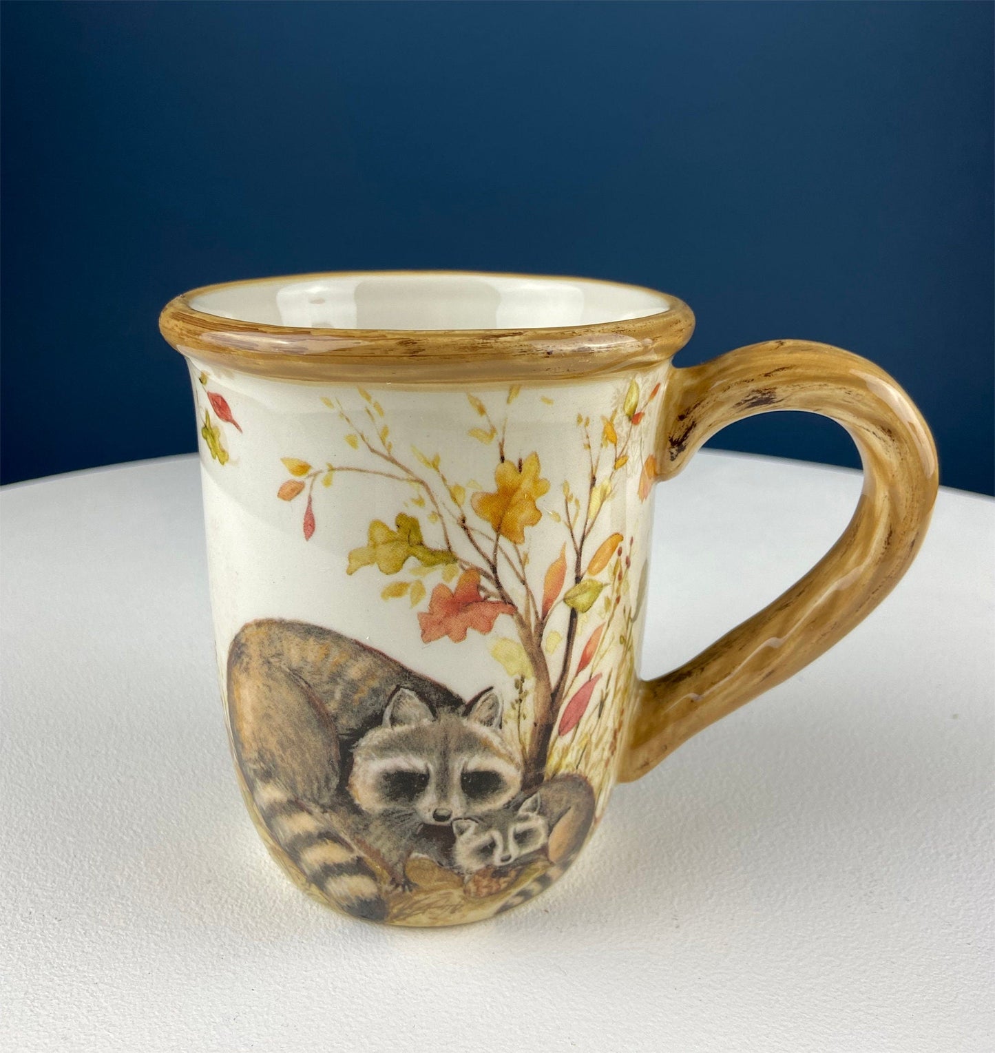 Ceramic mug with raccoon and autumn leaves design on a white surface with blue background