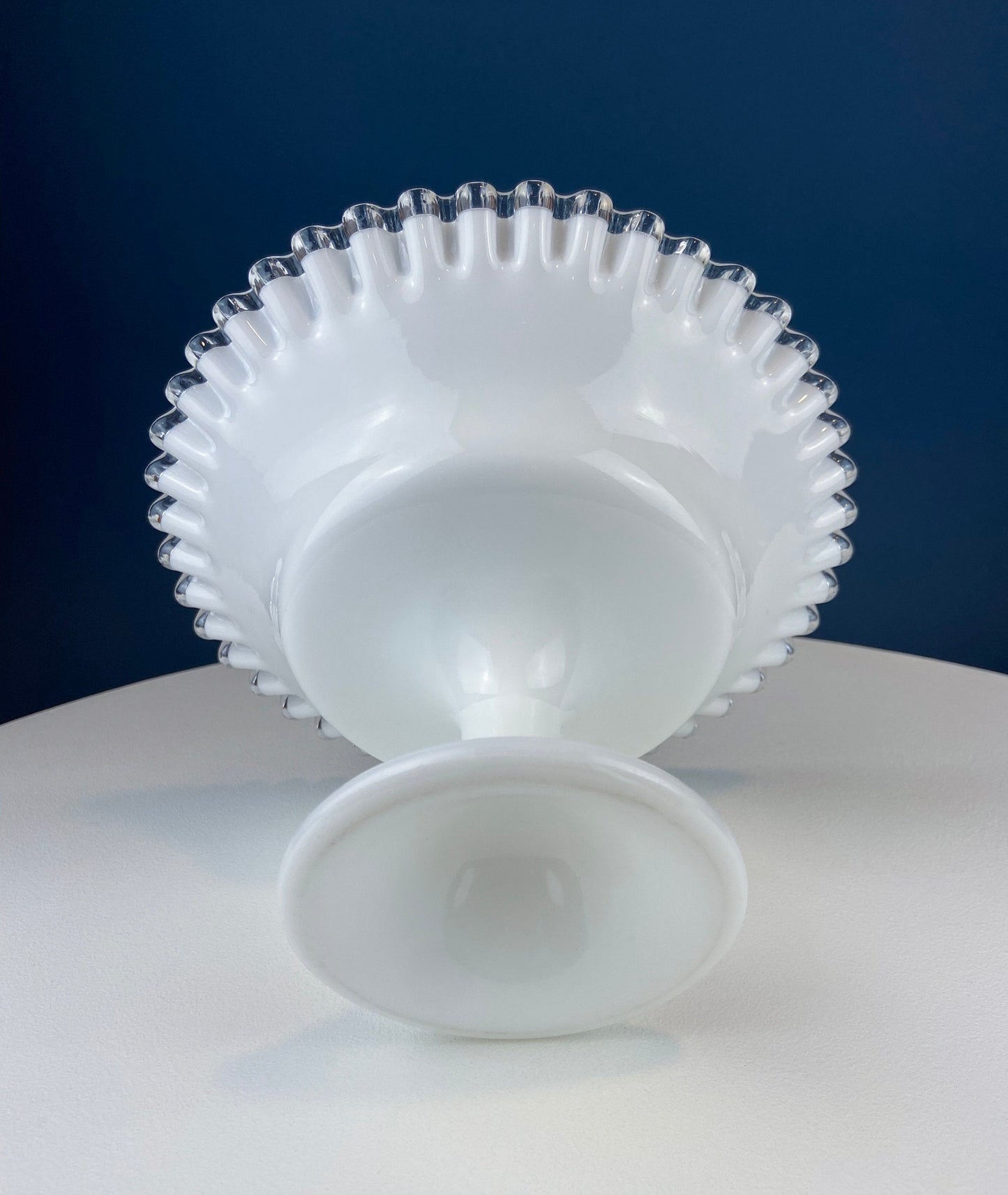 White glass footed bowl with textured rim on a white surface against a blue background