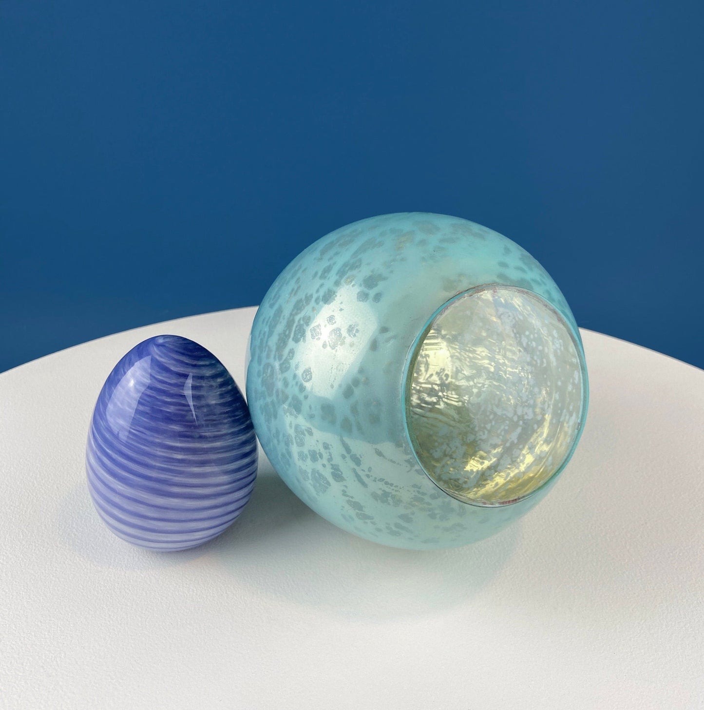 Two glass eggs, one blue and one green, on a white surface with a blue background
