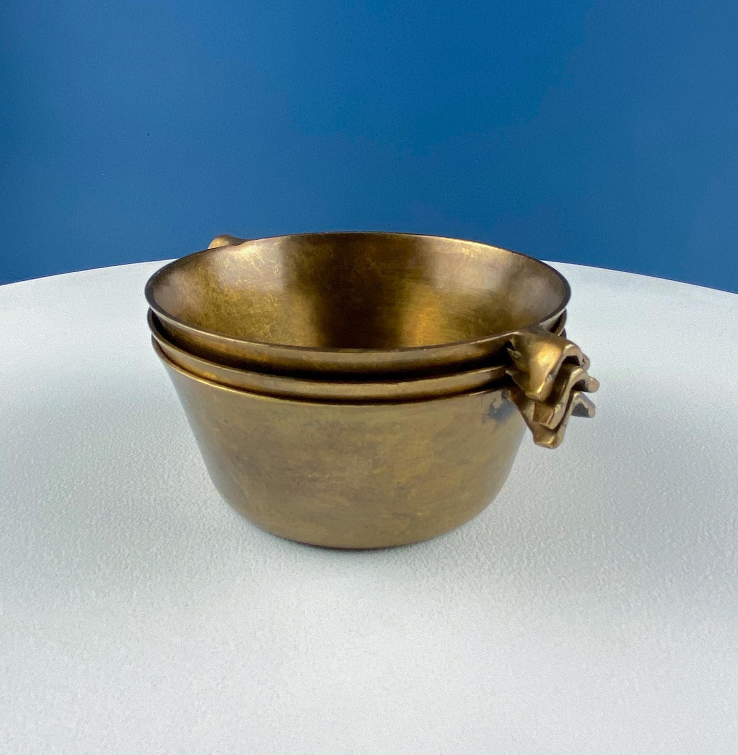 Stack of brass bowls with decorative handles on a white surface with a blue background
