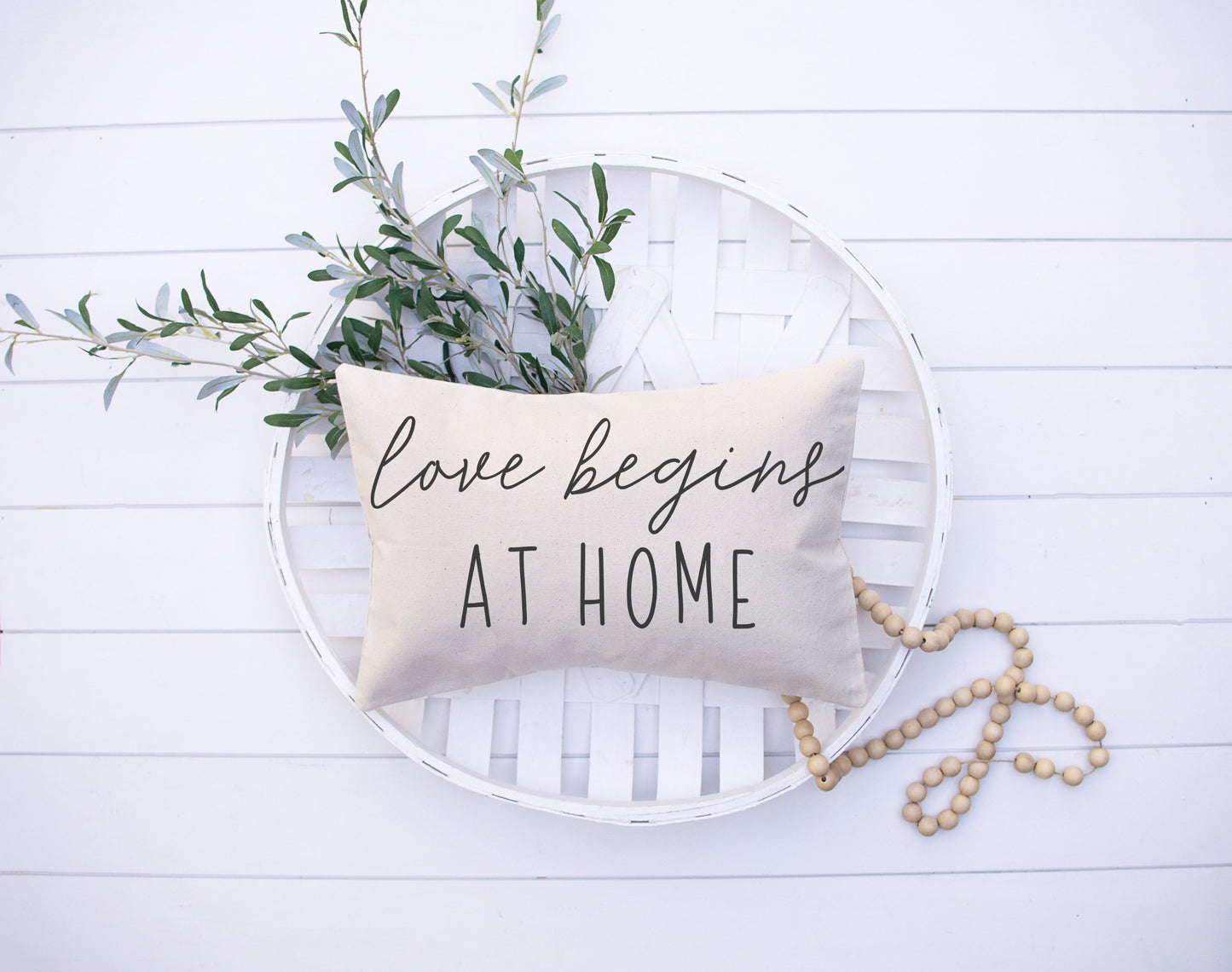 Love Begins At Home 12x20 inch Pillow Cover with oatmeal background and decorative greenery.
