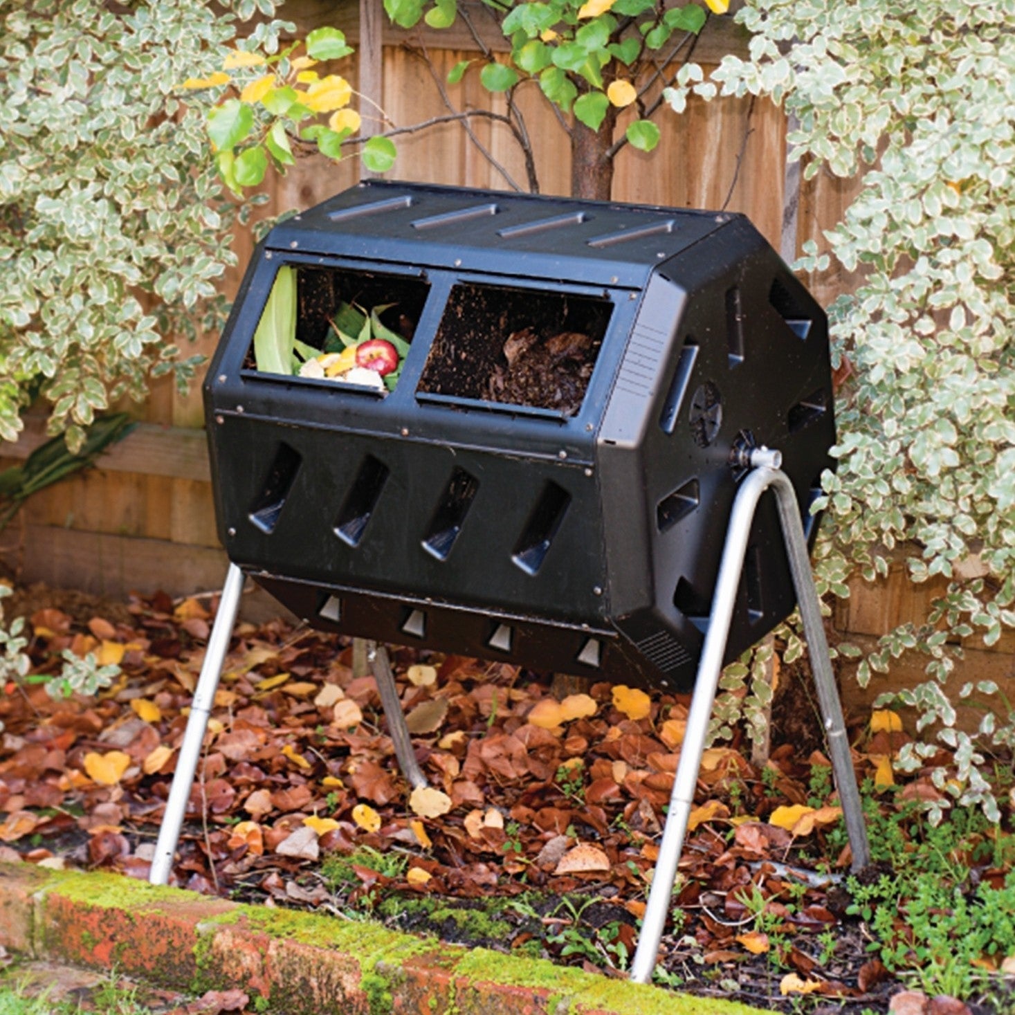 A black compost tumbler composter in a garden with greenery and fallen leaves.