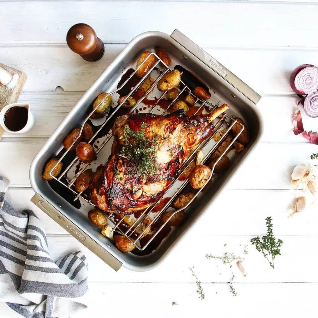 Roasting Pans with a roasted leg of lamb and golden potatoes on a wire rack, herbs sprinkled on top.