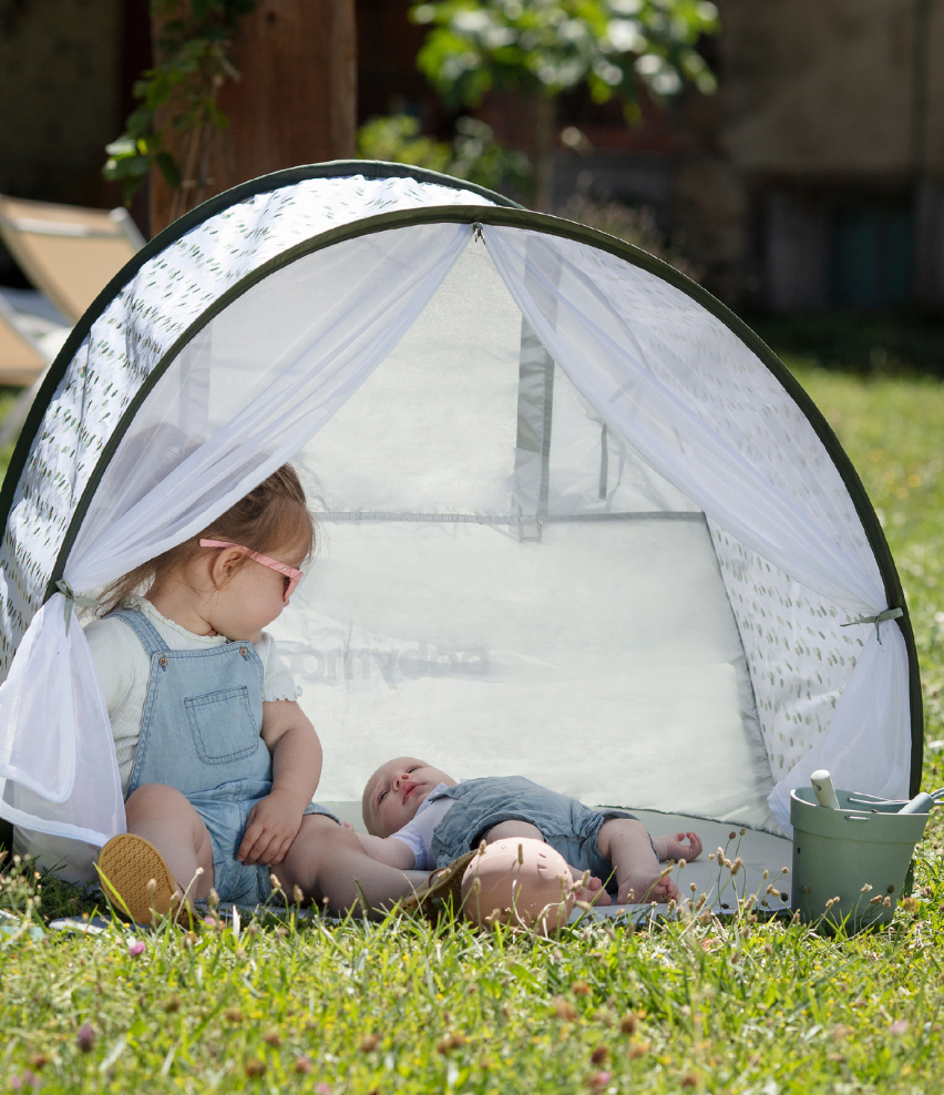 A young girl relaxing with a baby inside a Baby Beach Tent on a sunny day in a grassy area.