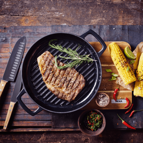 Griddles and Grill Pans with a grilled steak, corn, and fresh herbs on a rustic wooden table.