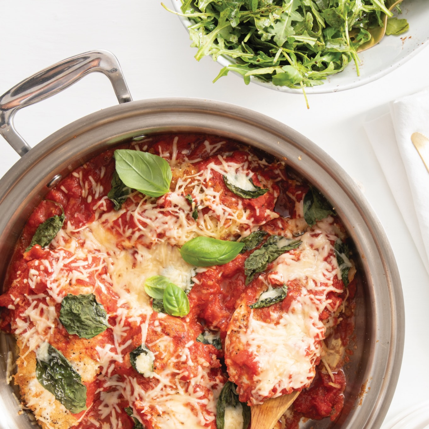 Sauté Pans with chicken Parmesan topped with cheese and basil, served with a side salad on a white background.