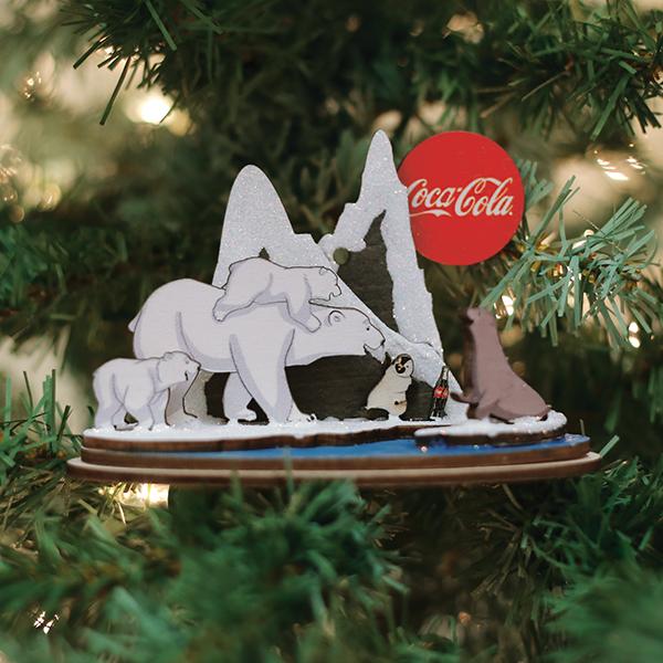 Decorative Christmas ornament with animals and Coca-Cola logo on a tree.