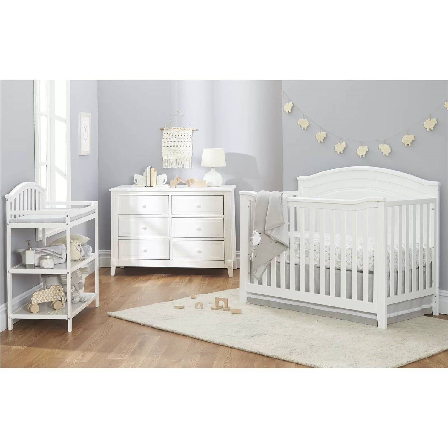 White nursery furniture set including crib, changing table, and dresser in a room with light gray walls and wooden flooring.