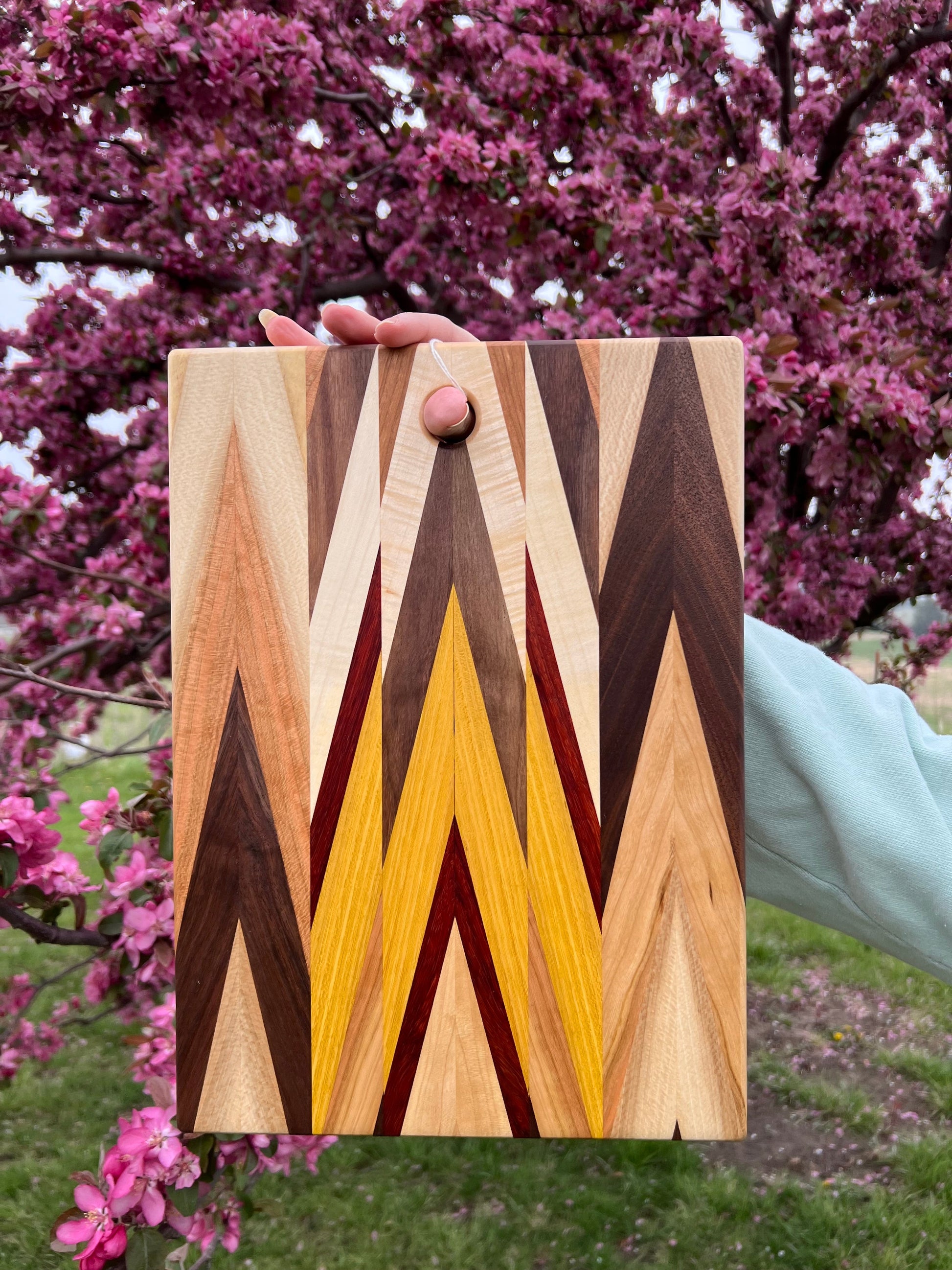 Wooden cutting board with a geometric pattern held in front of a flowering tree
