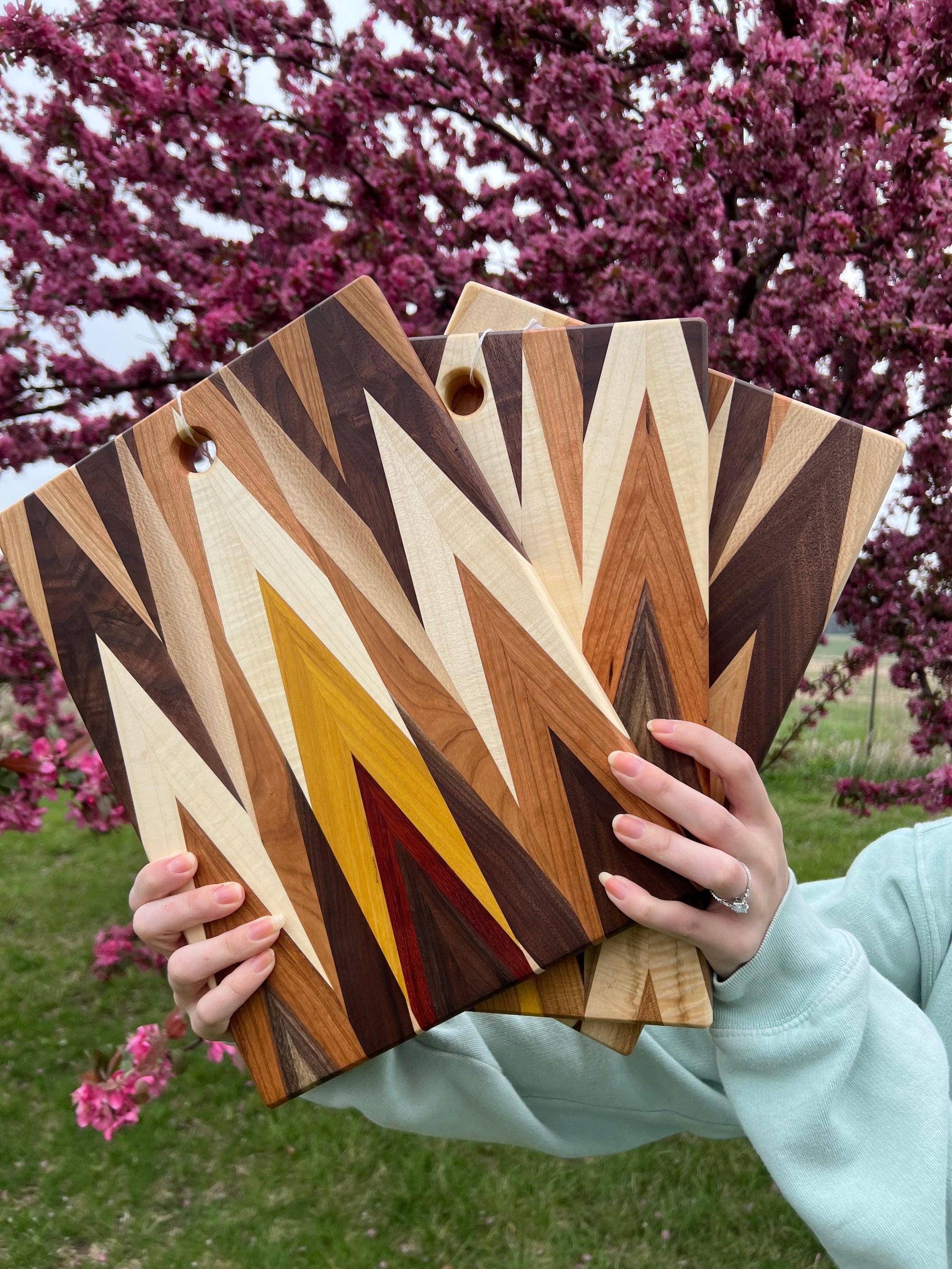 Three wooden cutting boards with a geometric pattern held by a person outdoors with cherry blossoms in the background.