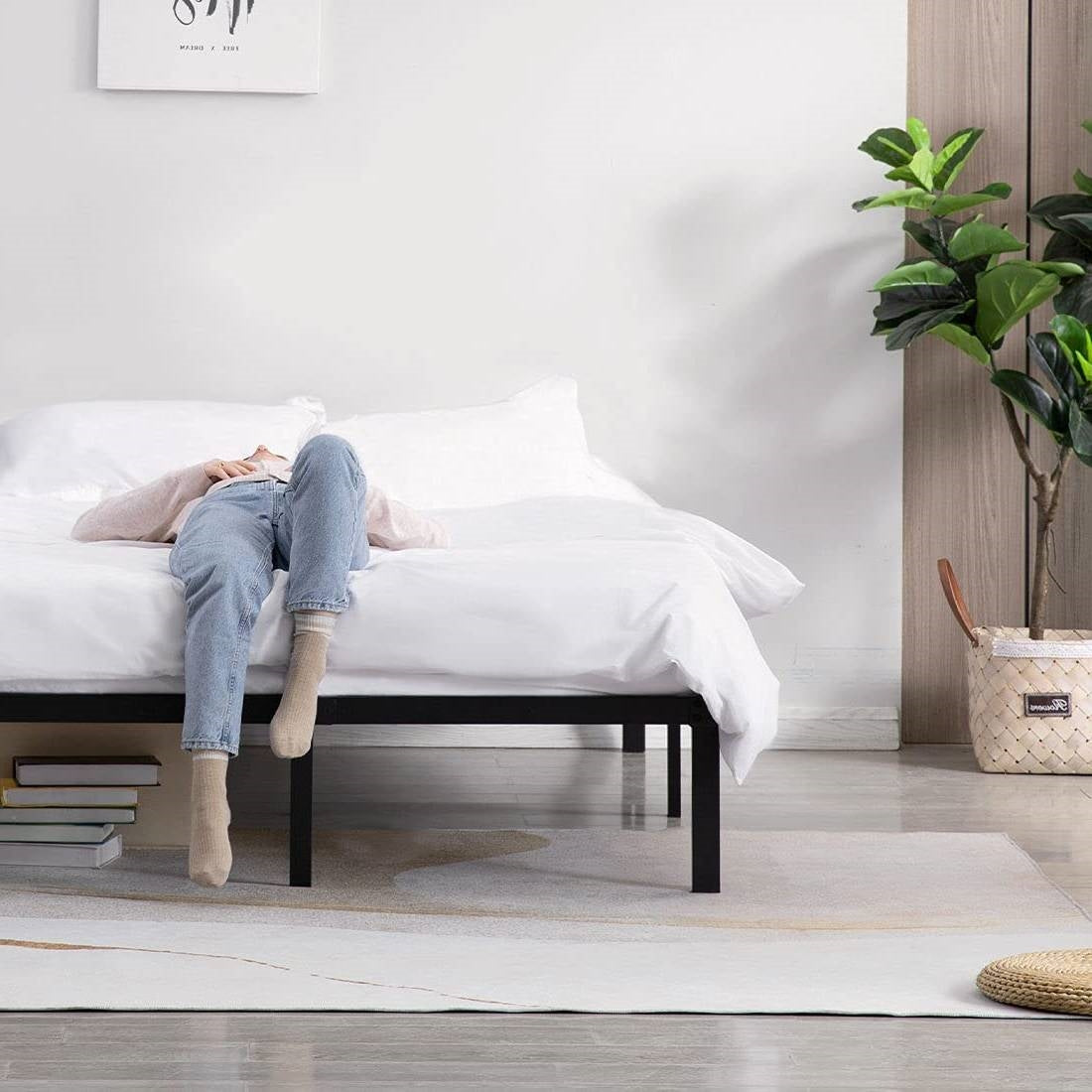 Person lying on a white bed in a minimalistic room with a plant and books.