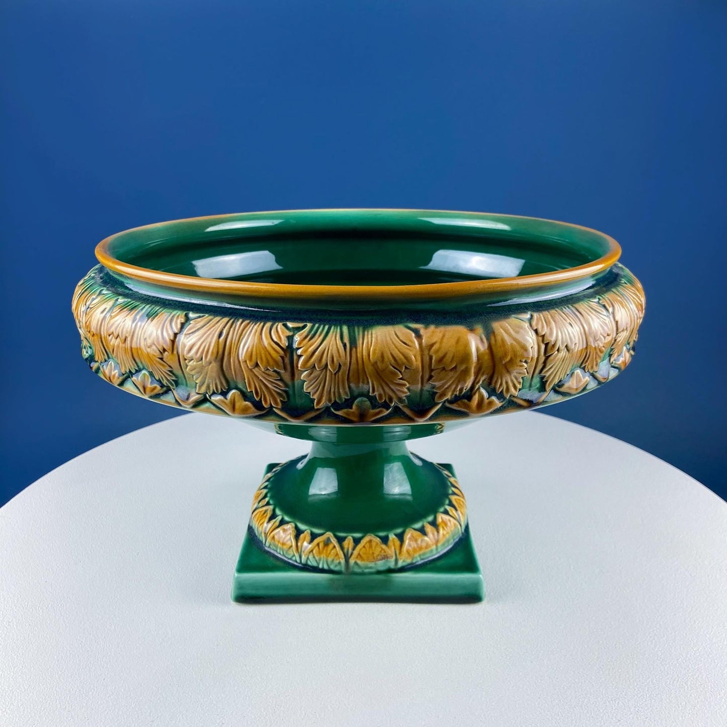 Decorative green and gold ceramic bowl on a white pedestal against a blue background