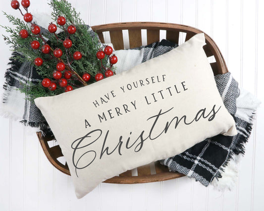 Merry Little Christmas Pillow Cover 12x20 inch on a basket with holiday decorations and cozy blankets.