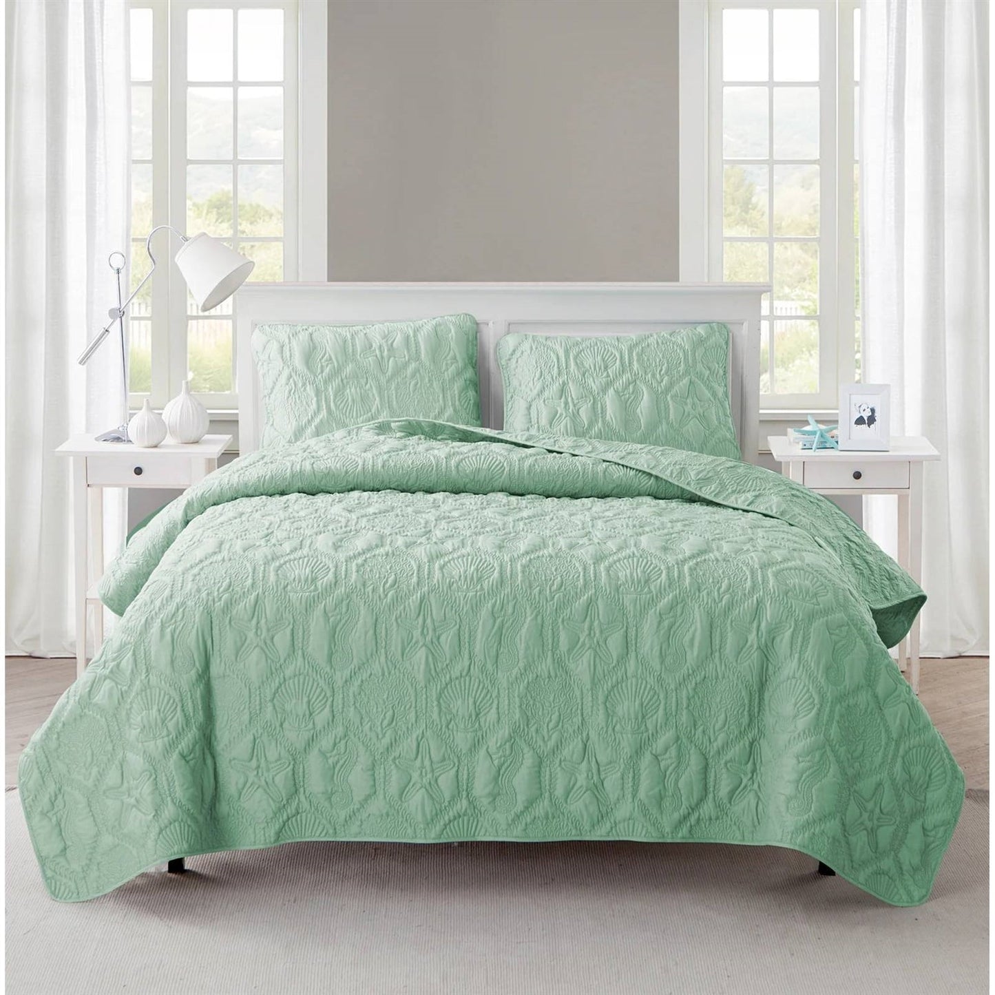Green quilt set on a bed in a bright bedroom with large windows.