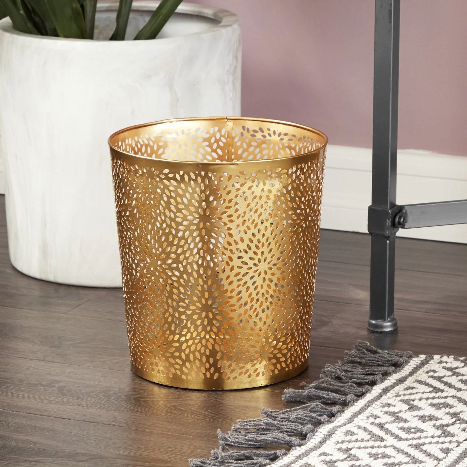 Gold decorative trash can on a wooden floor with a plant and rug in the background
