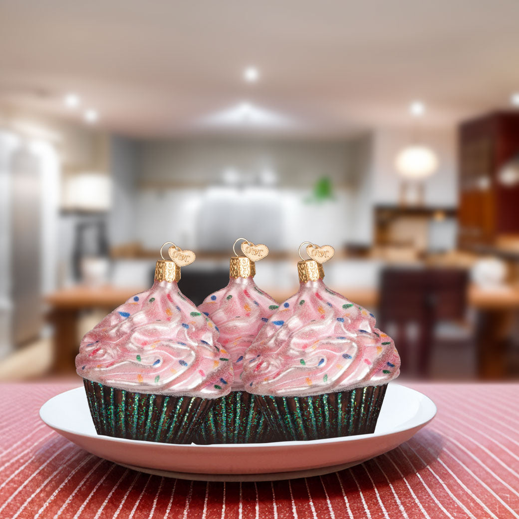 Three pink frosted cupcakes with sprinkles on a plate in a blurred kitchen setting