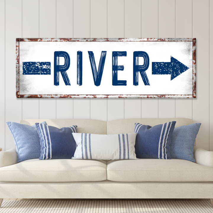 Decorative sign with 'RIVER' and arrows on a wall above a sofa with blue and white pillows.