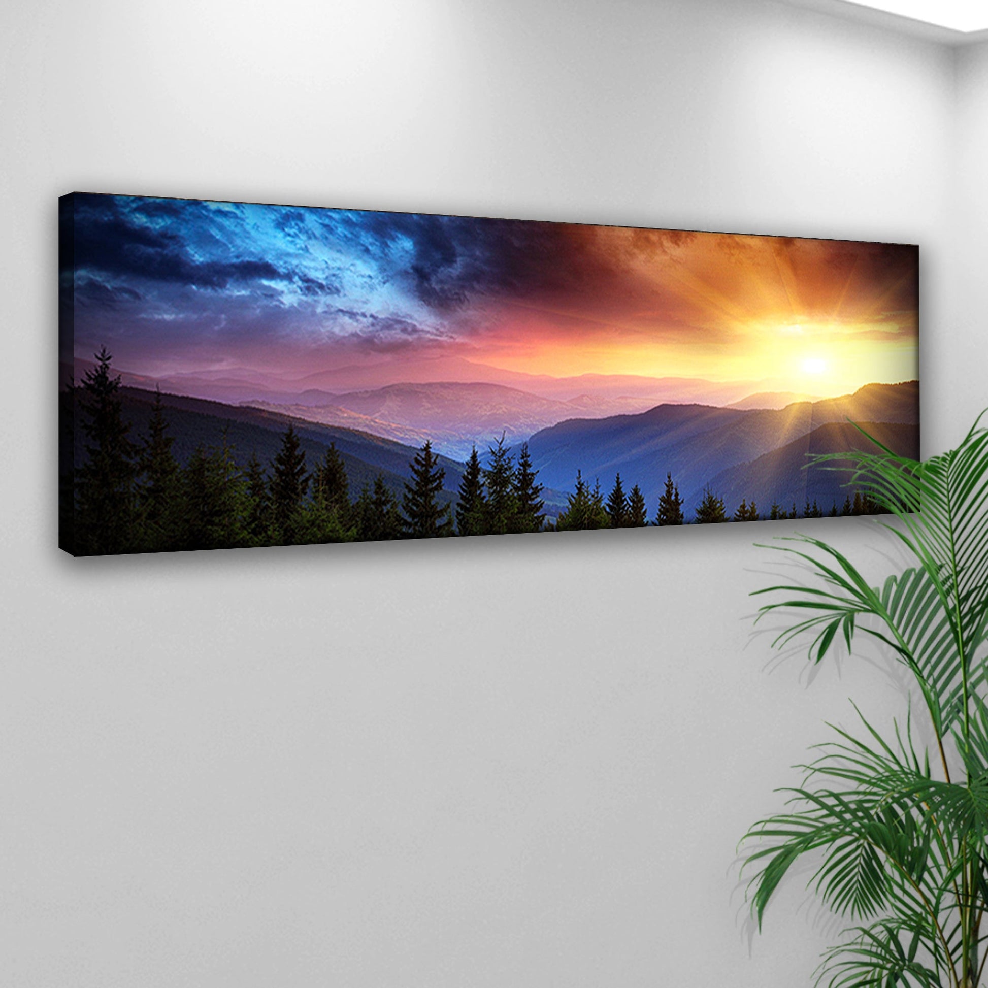 Panoramic landscape print of a sunset over mountains on a white wall with a plant in the corner.