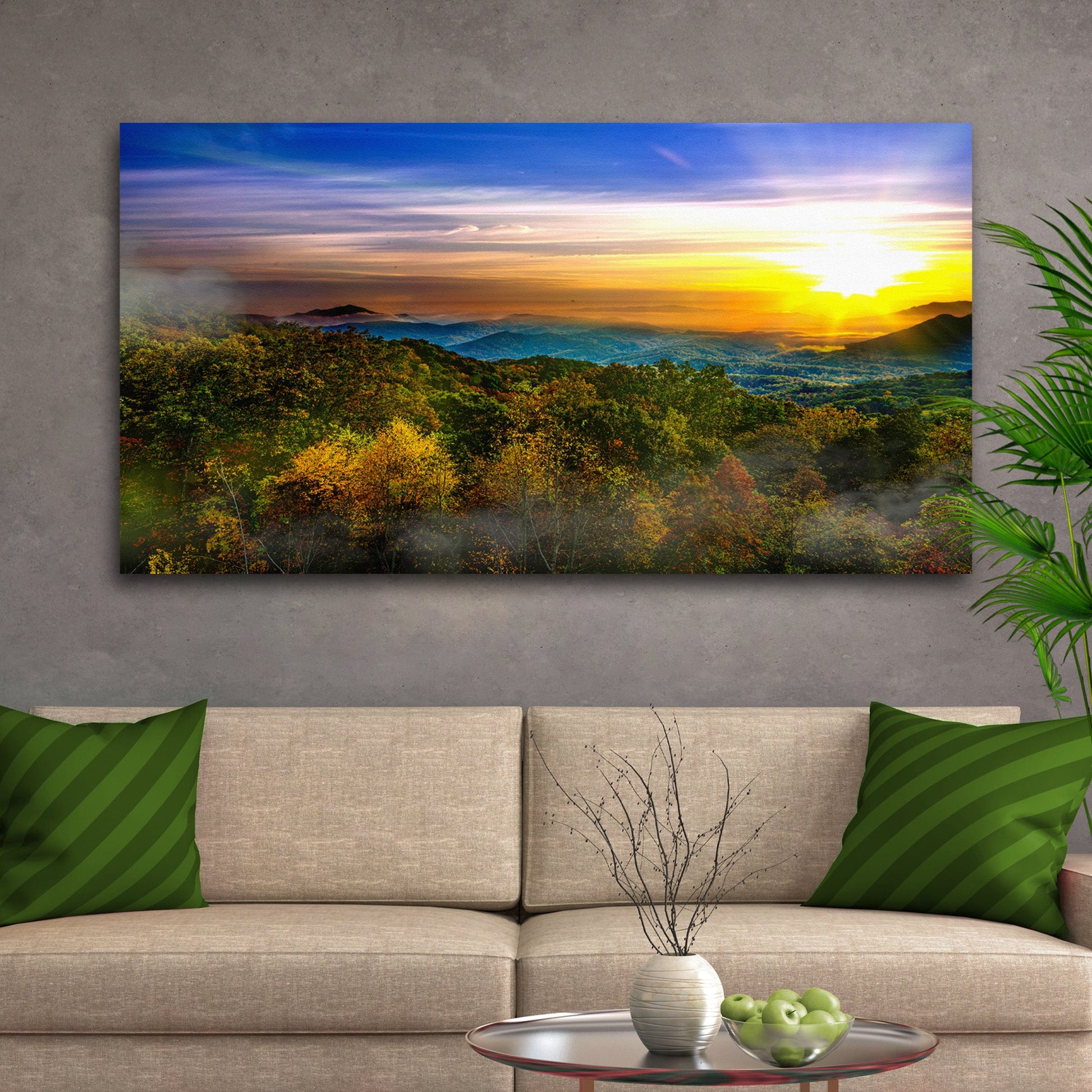Sofa with green pillows and a scenic print of a sunset over trees on a gray wall.