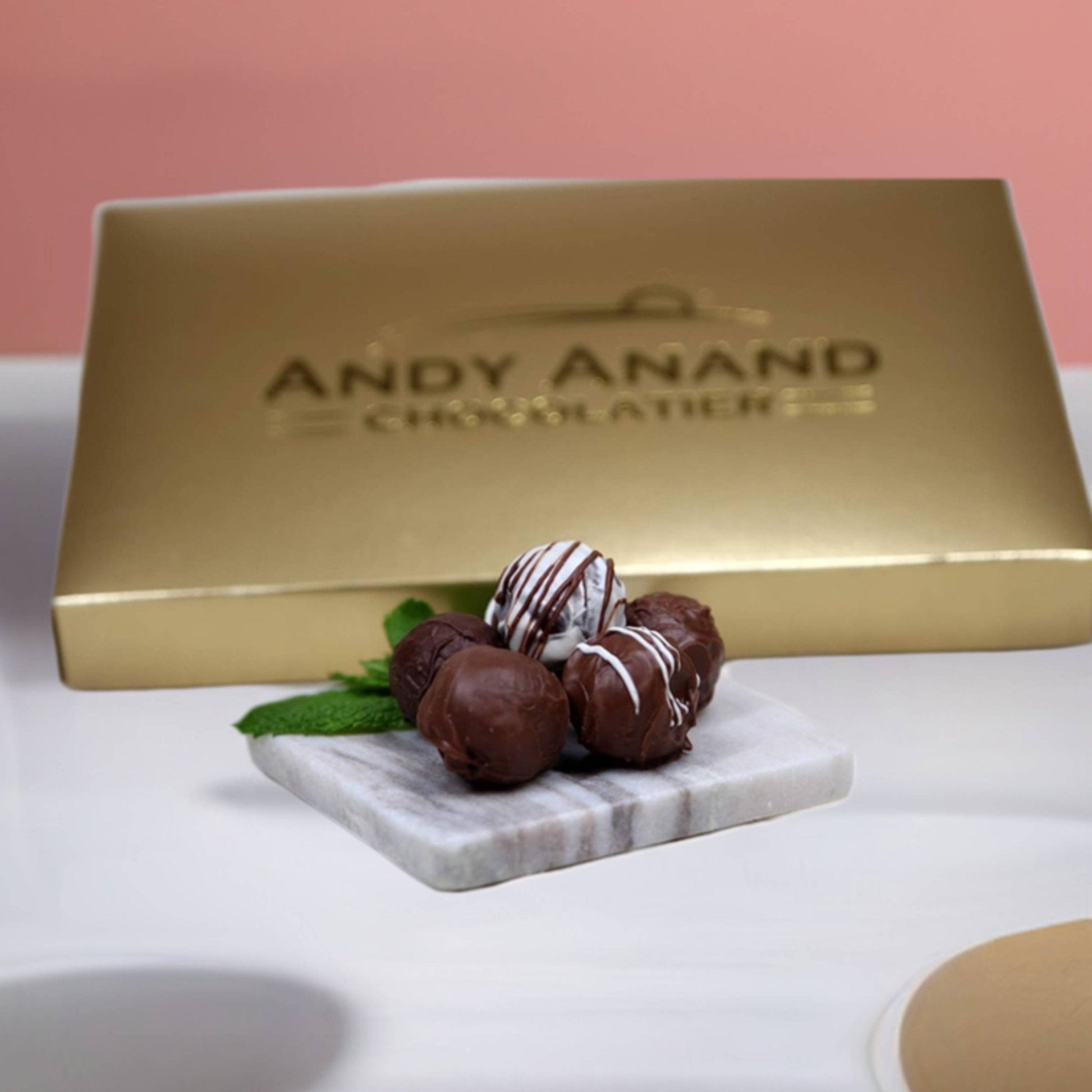 Gold chocolate box with 'Andy Anand Chocolates' branding, accompanied by chocolate truffles on a marble board.