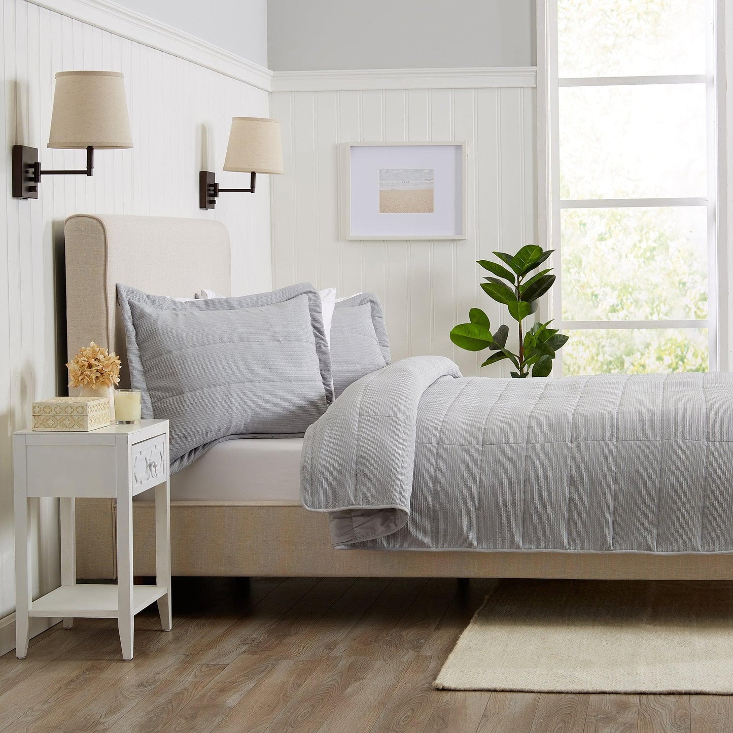 Neatly made bed with gray bedding in a bright bedroom with a side table and plant.