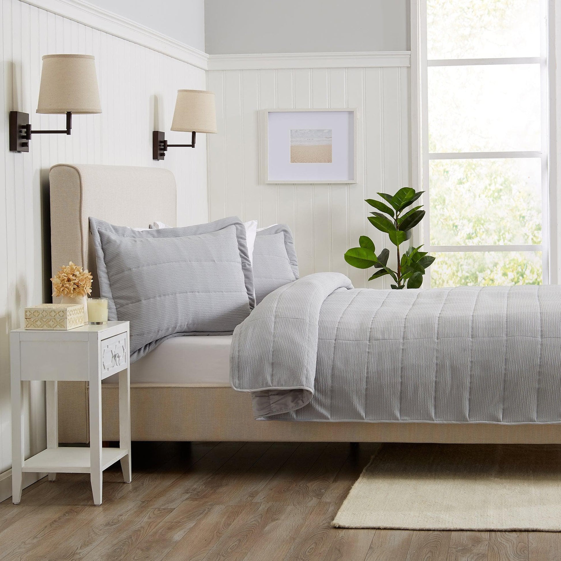 Neatly made bed with gray bedding in a bright bedroom with a side table and plant.