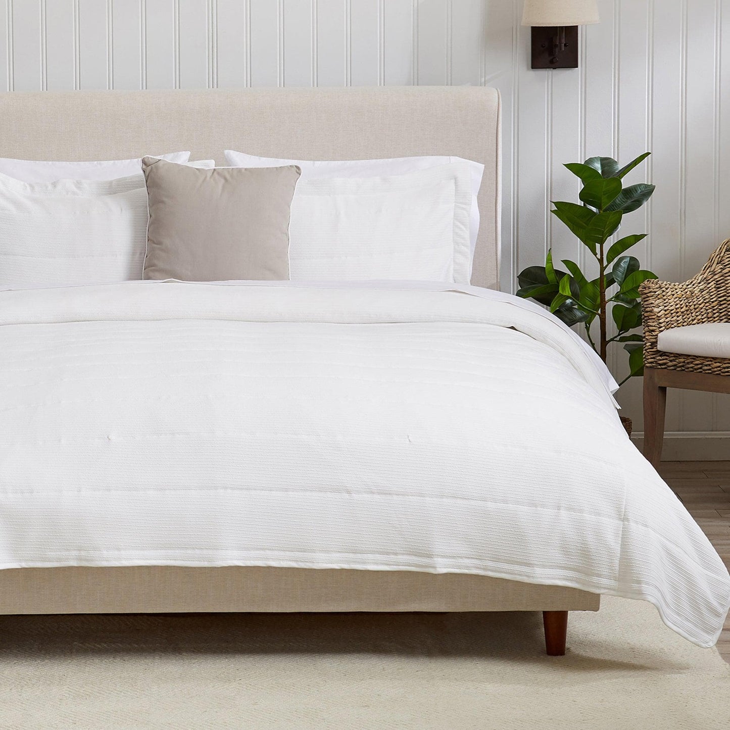 Neatly made bed with white bedding and a beige headboard in a room with a plant and chair.
