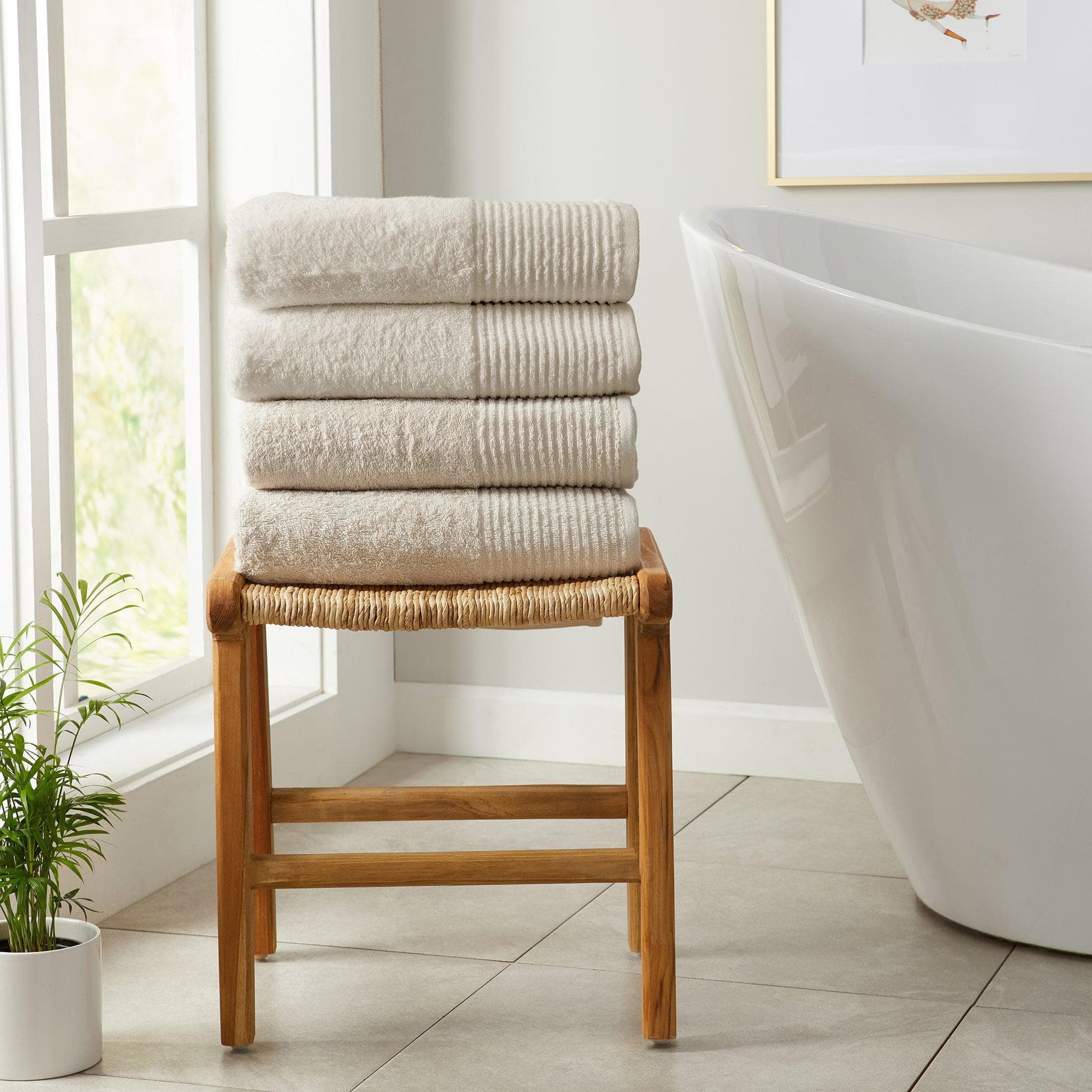 Stack of towels on a wooden stool in a bathroom with a bathtub and window in the background.