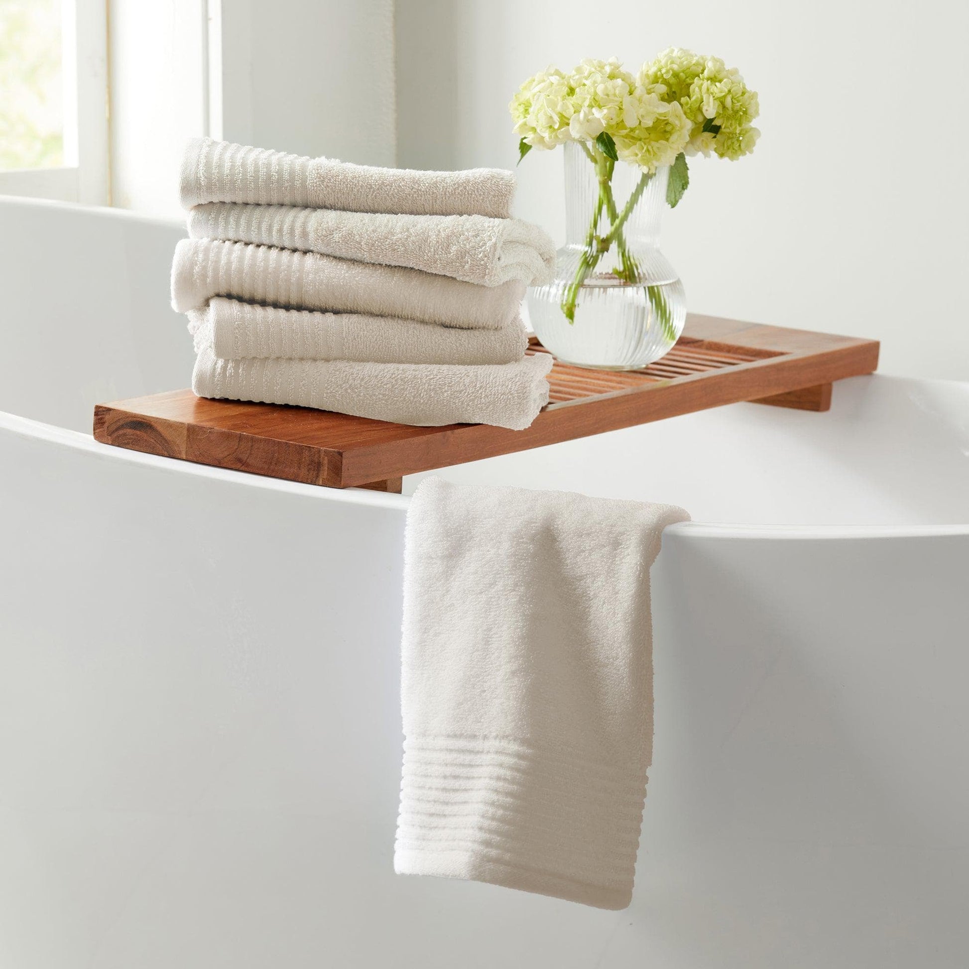 Stack of white towels on a wooden tray over a bathtub with a vase of flowers.