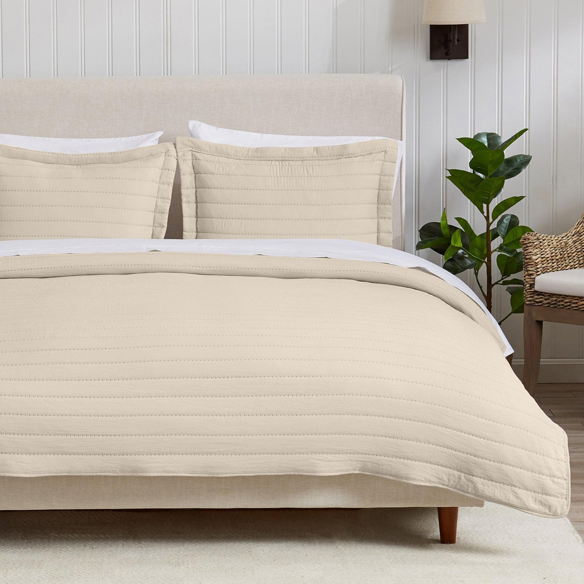 Beige quilted comforter set on a bed with pillows and a plant in the background.