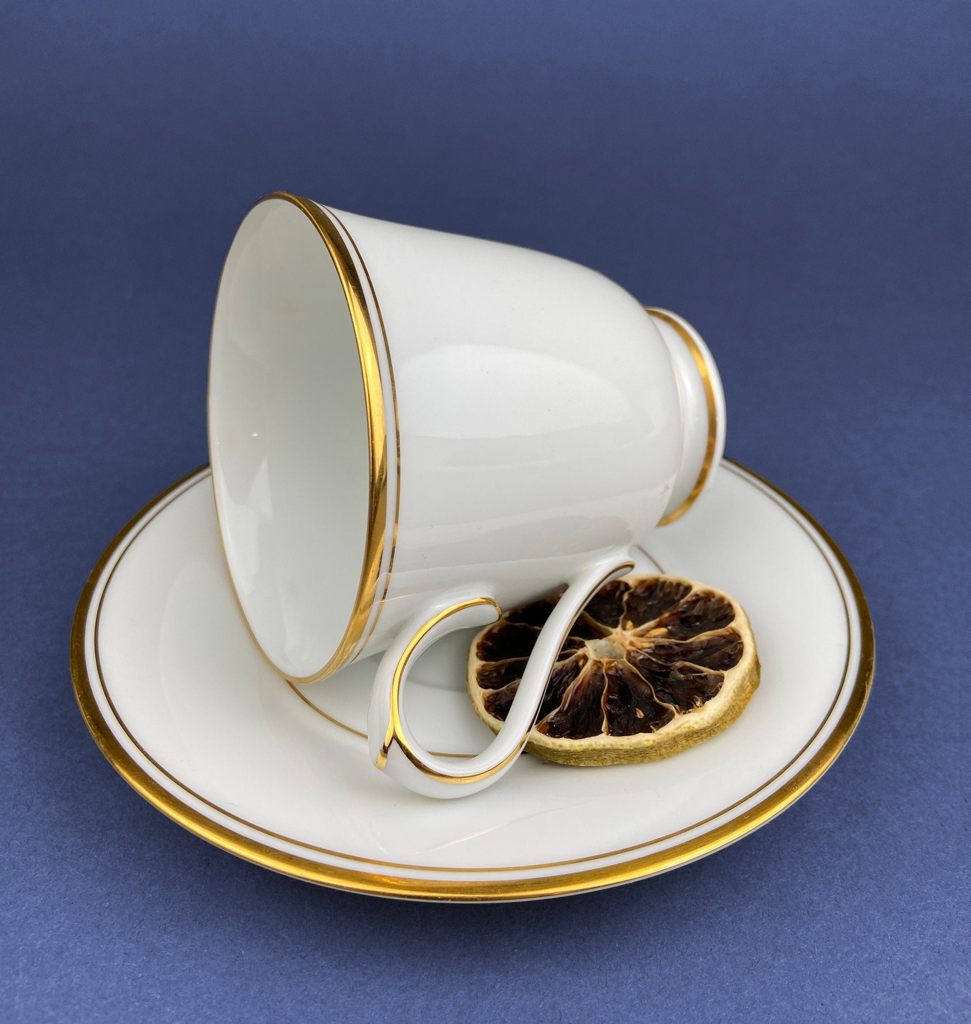 White teacup with gold trim on a matching saucer, containing dried lemon slices against a blue background.