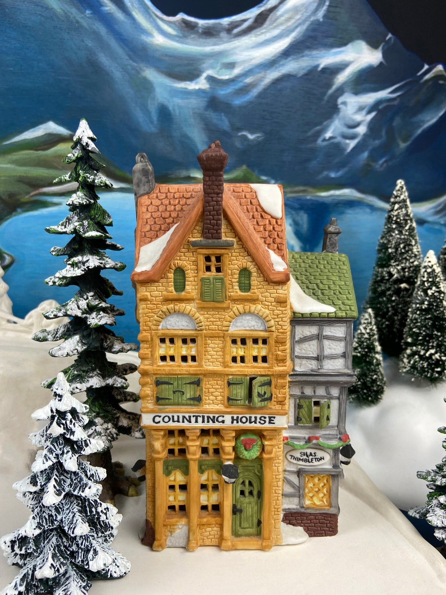 Decorative model of a 'Counting House' with snow-covered trees and mountains in the background.