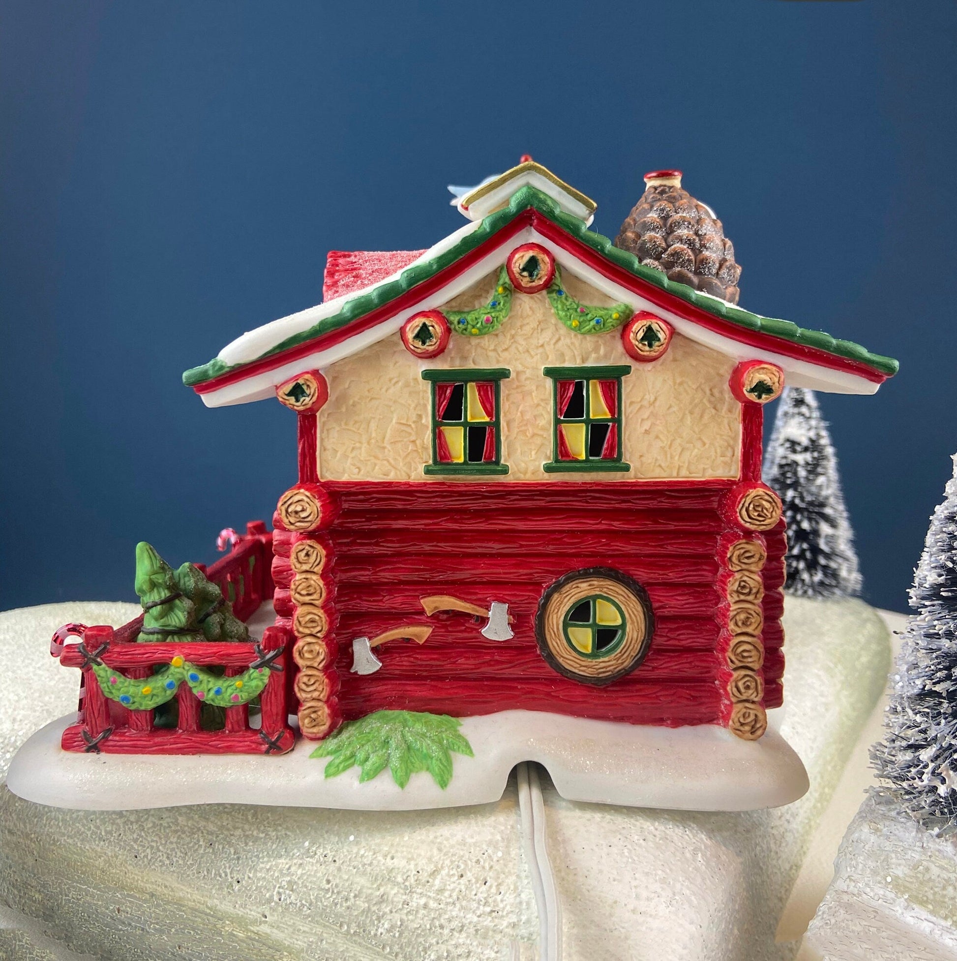 Decorative log cabin ornament with a snowy base and Christmas trees in the background.
