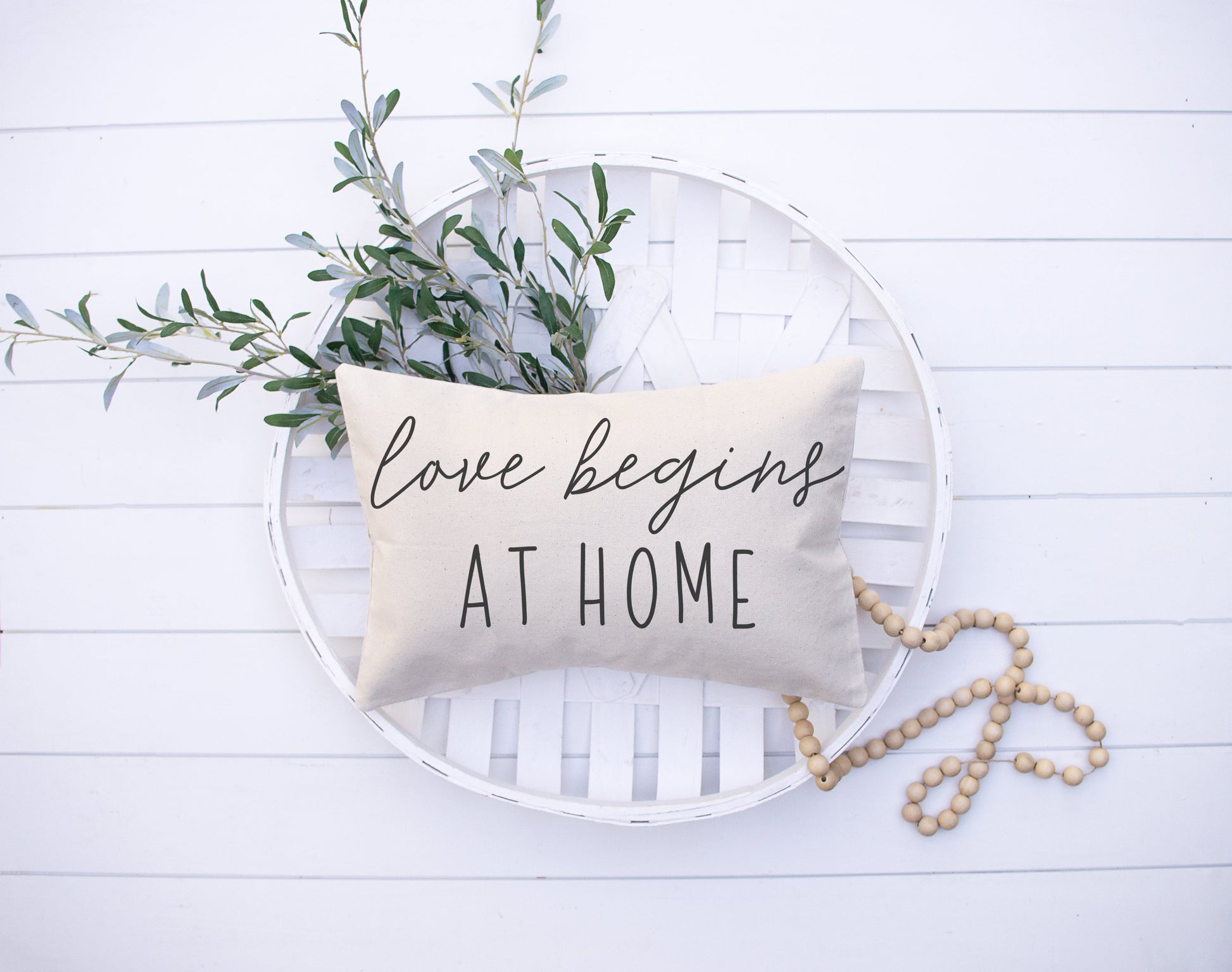 Love Begins At Home 12x20 inch Pillow Cover with oatmeal background and decorative greenery.