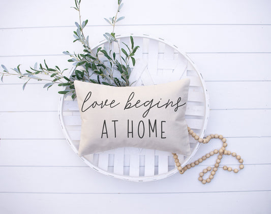 Love Begins At Home 12x20 inch Pillow Cover with oatmeal background and decorative greenery.