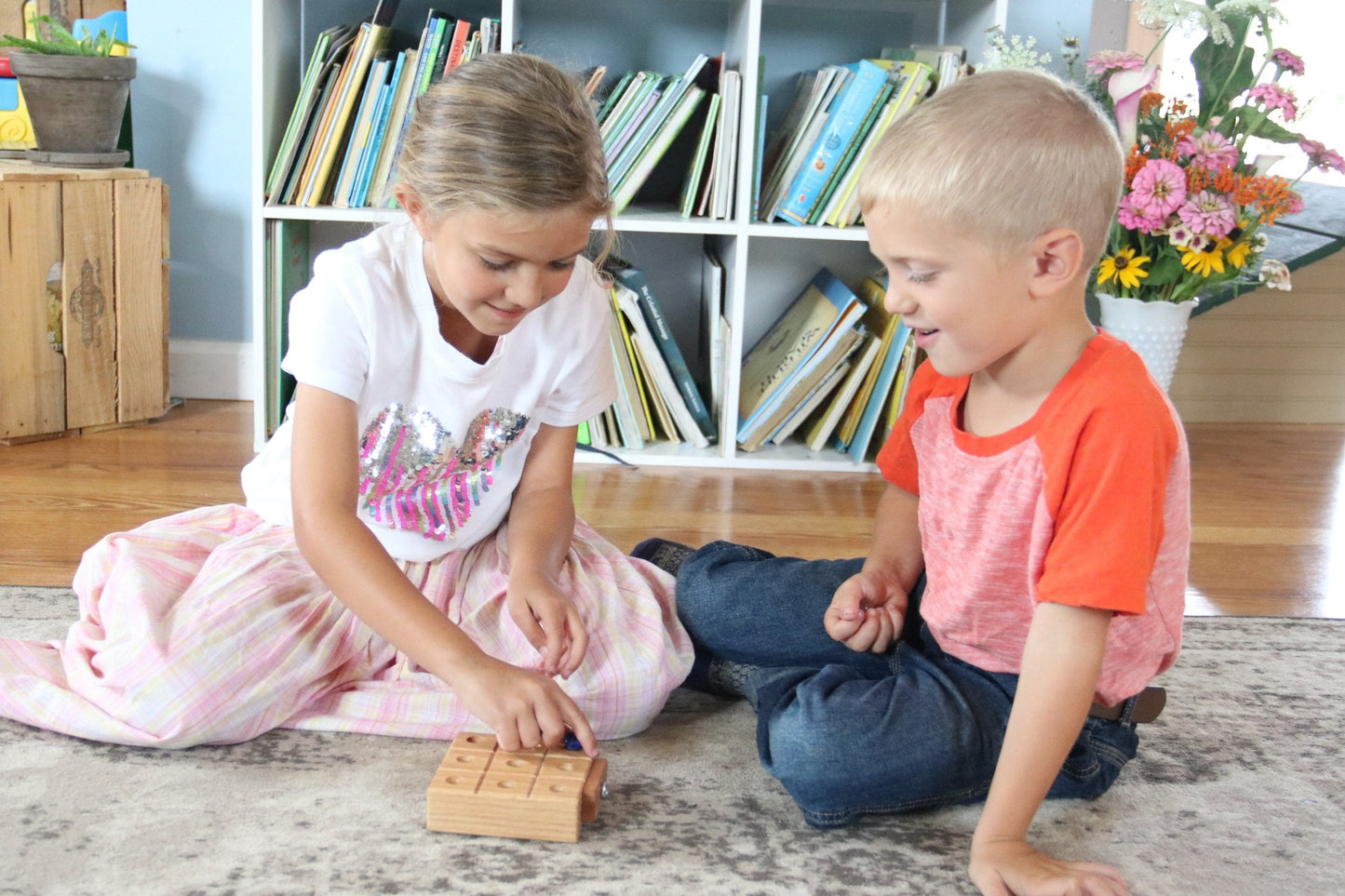 Two children playing with wooden tic-tac-toe on a carpeted floor in a room with bookshelves and flowers.