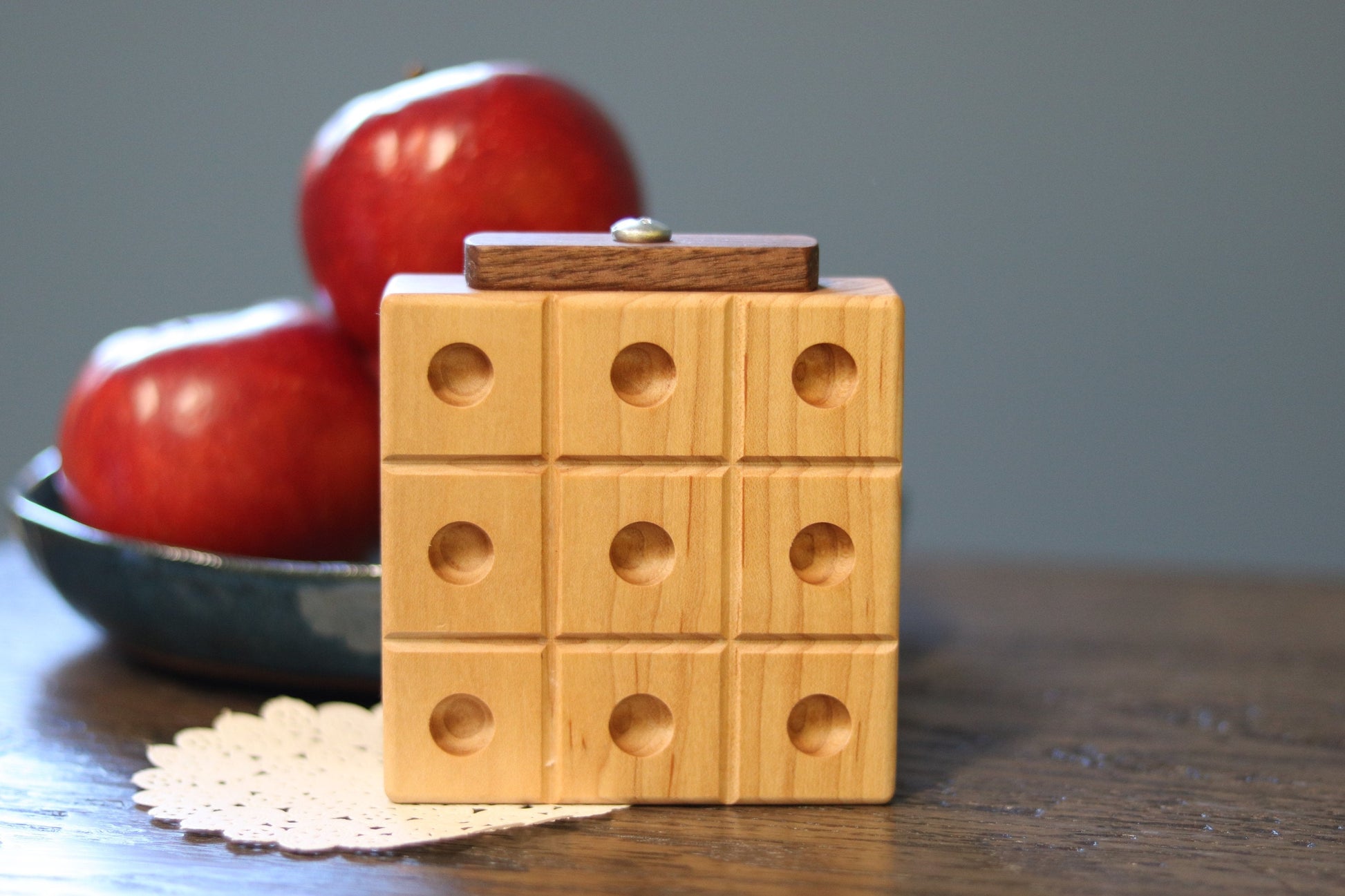 Wooden tic-tac-toe box with circular holes on a table with apples in the background