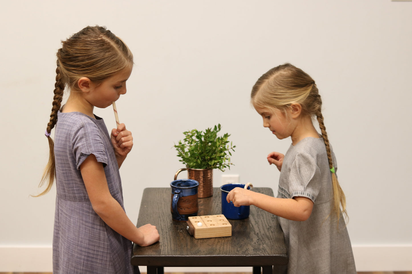 Two young girls playing with wooden toys at a table against a plain wall.