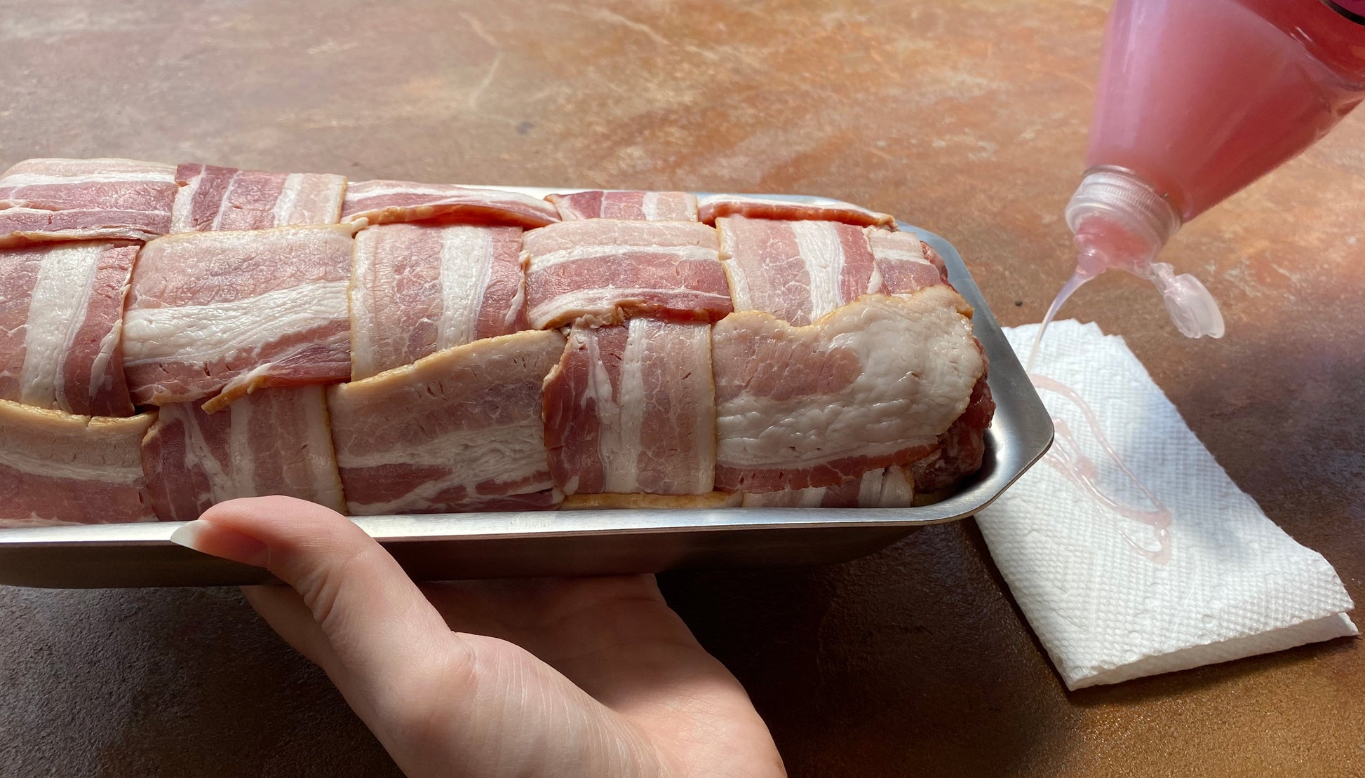 Bacon-wrapped meat on a tray with a hand holding a bottle of liquid for seasoning or glazing.