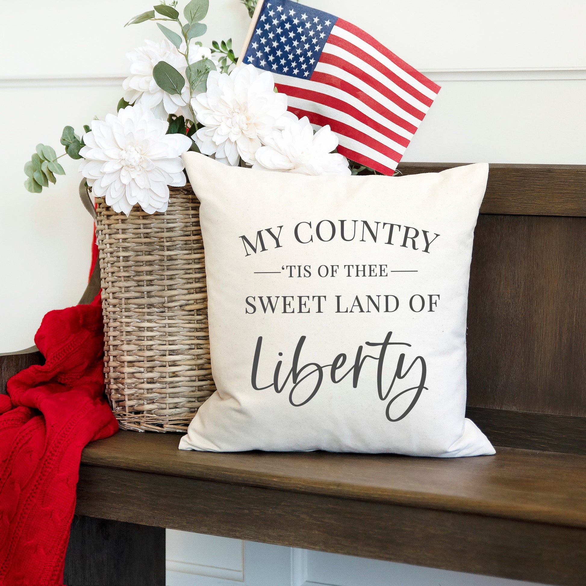 My Country Tis of Thee Summer Pillow Cover 18x18 inch displayed on a bench with flowers and American flag.