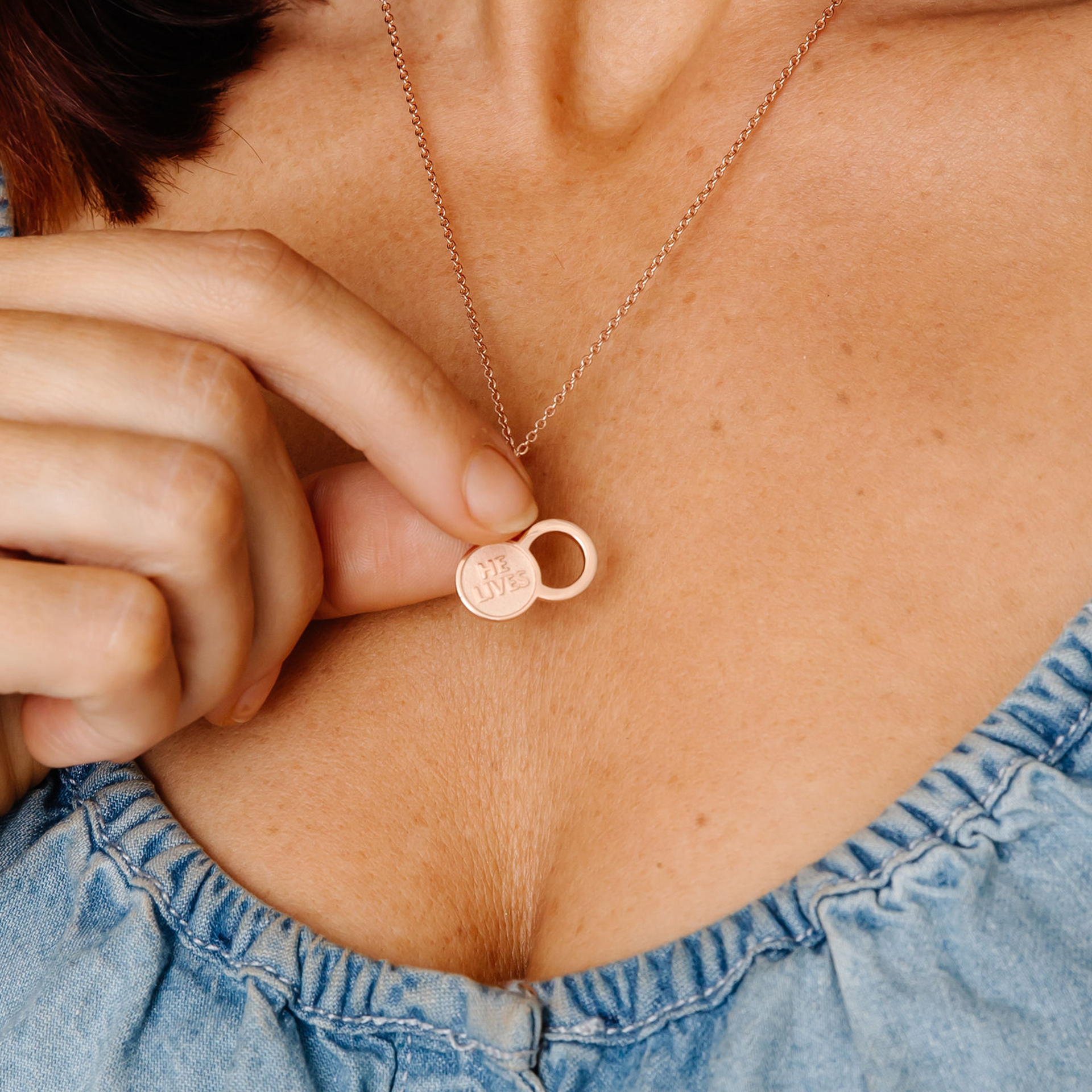 A close-up of a woman holding the Empty Tomb Pendant Necklace, showcasing the 'He Lives' inscription.