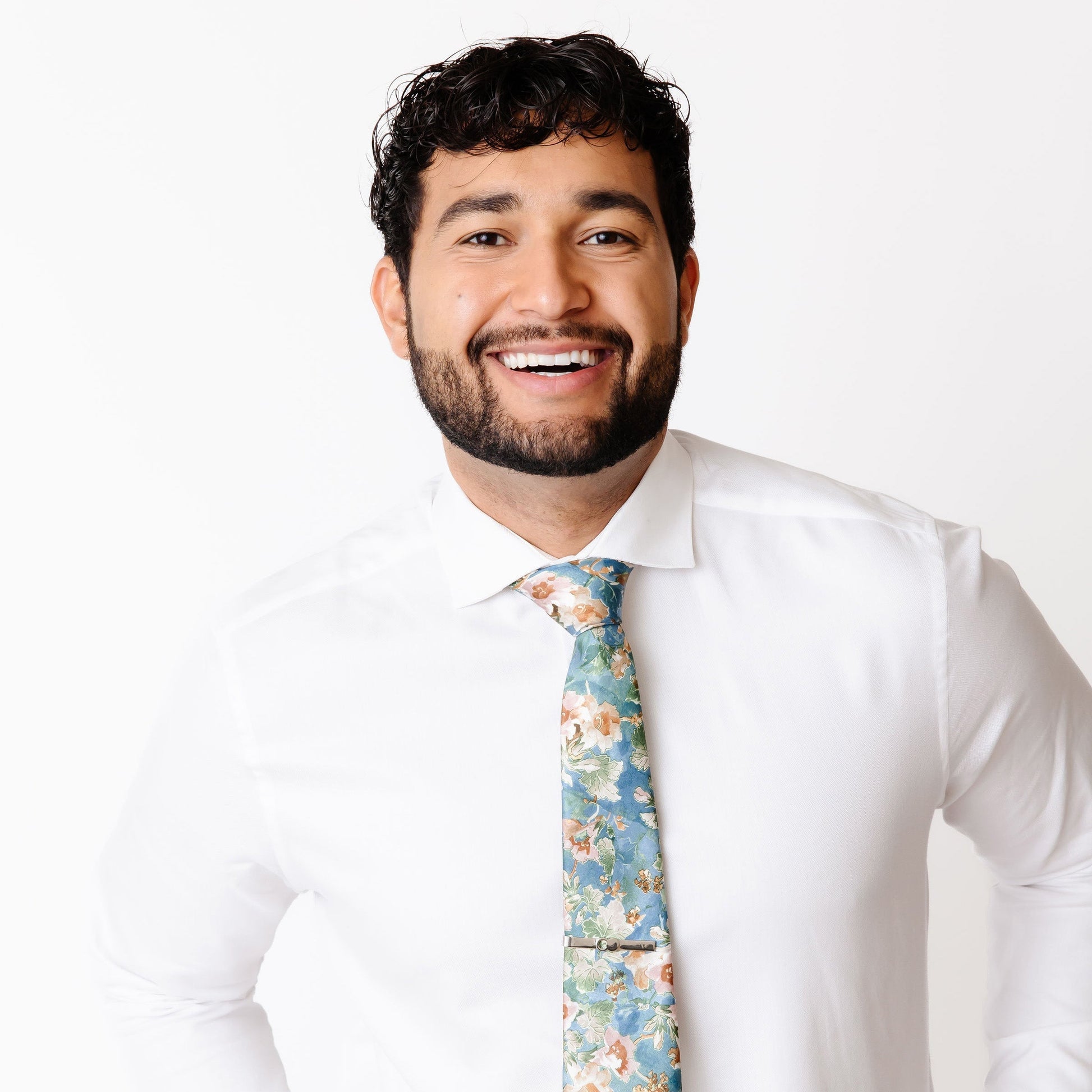 A man smiling and wearing a floral tie with a white shirt, showcasing a modern and stylish look.