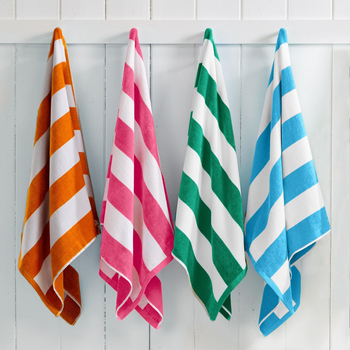 Four striped beach towels in orange, pink, green, and blue hanging on a white wooden panel wall.
